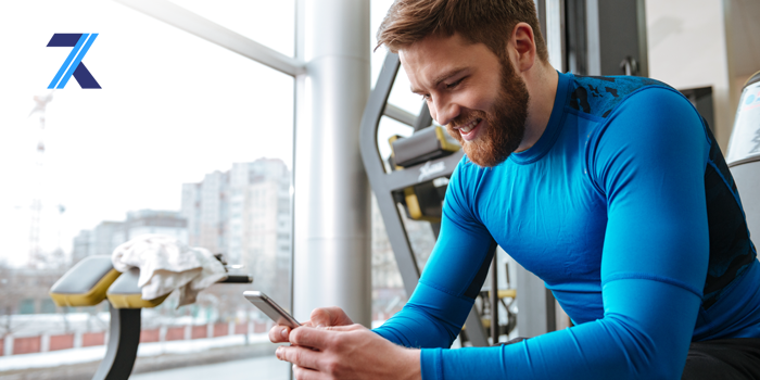 A bearded man wearing a blue athletic top sitting in a modern gym while looking down and smiling at the smartphone he is holding.